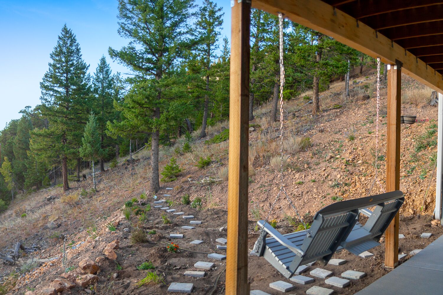 Ett takförsett utomhusområde med sittplatser och en bänk, omgivet av en träpergola, med utsikt över en bergig sluttning med träd.