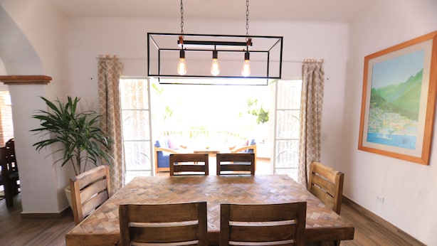 Uma sala de jantar com uma mesa e cadeiras de madeira em padrão herringbone, um lustre e um vaso de planta.
