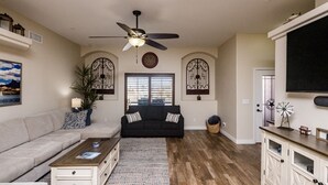 Ein Wohnzimmer mit einer Couch, einem Couchtisch und einem Deckenventilator. Ein Fenster mit Jalousien, eine Tür und ein an der Wand befestigter Fernseher sind ebenfalls vorhanden.