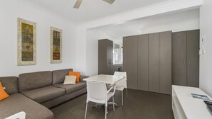 Un salon moderne avec un canapé gris, des chaises blanches et une petite table. Deux œuvres d’art ornent le mur, et un coin cuisine est aménagé avec des meubles gris.