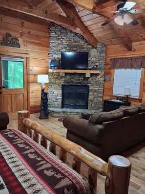 A log cabin interior with a stone fireplace, wooden ceiling beams, a television, and a cozy seating area.