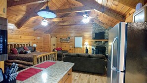 A cozy cabin interior with a stone fireplace, wooden ceiling, a kitchen area with a refrigerator, and a seating area with a couch and a table. - Newly updated cabin, 10 min to pkwy in Pigeon Forge. Screen porch, hot tub,WiFi. (Sevierville)