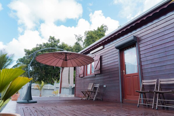 Un bâtiment en bois avec une porte rouge, un patio couvert avec une table et des chaises, et un grand parasol.