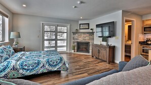 A spacious living room with a large bed, a fireplace, a TV mounted on the wall, and a view of the outdoors through a sliding glass door.