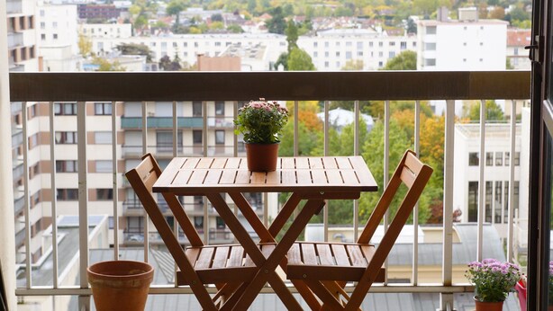 A balcony with a wooden table and chairs, a potted plant, and a view of a cityscape with buildings and trees. - Studio l'Orchidée Mobility lease (1 to 10 months) (Rueil-Malmaison)