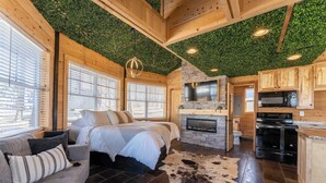 A cozy log cabin interior with a bed, sofa, fireplace, kitchen, and a ceiling with green foliage.