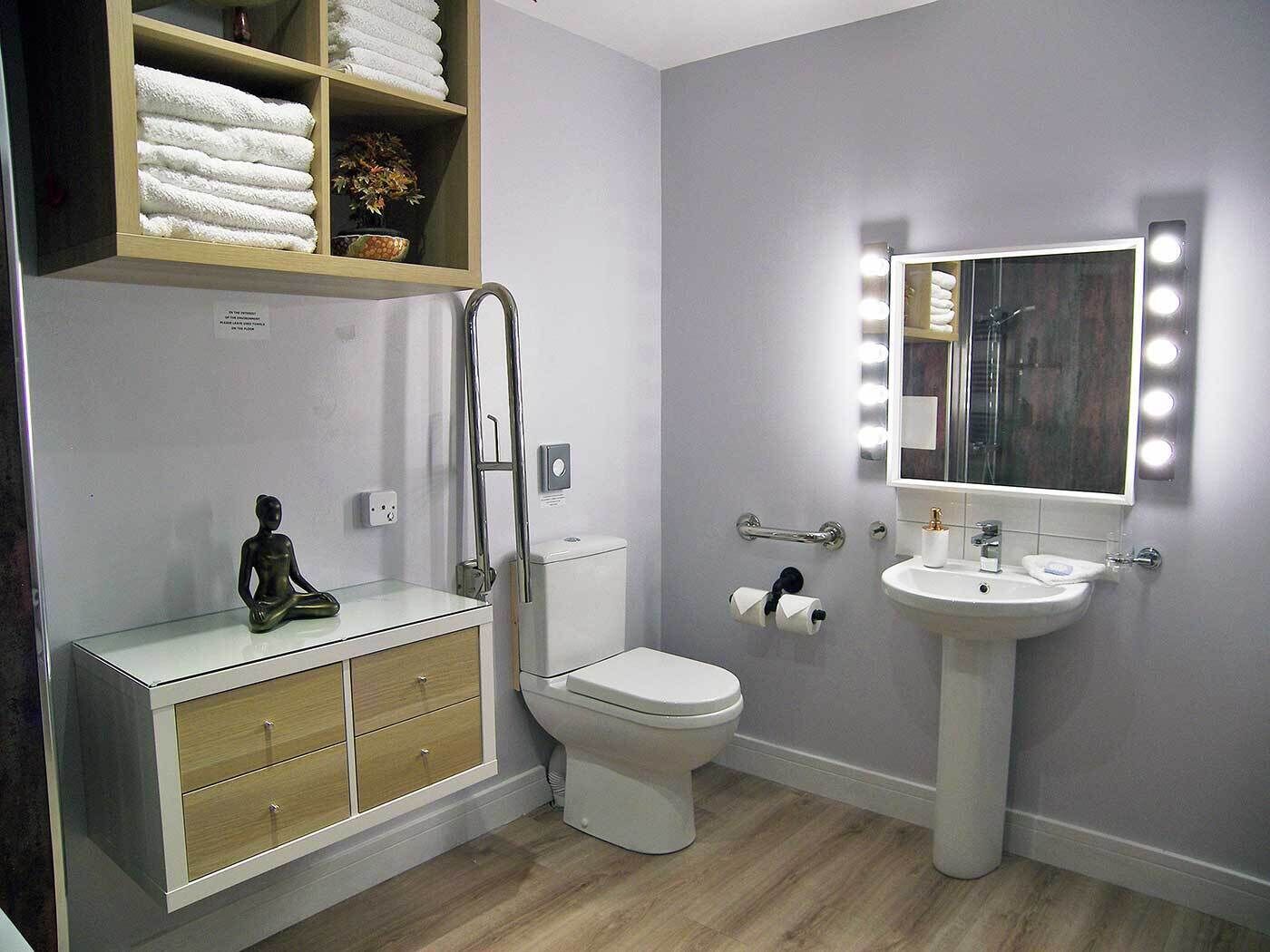 A modern bathroom with a toilet, sink, vanity with drawers, a shelf with towels, and a mirror with lights.
