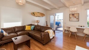 A spacious living room with a brown leather sofa, a coffee table, and a dining area with a white table and chairs. A surfboard and a guitar are visible on the wall, and there is a large window providing a view of the outdoors.