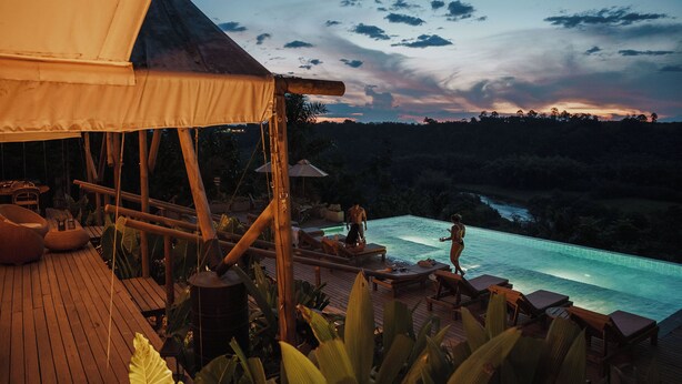 Terraza de madera con sillones de descanso y piscina, rodeada de plantas tropicales.