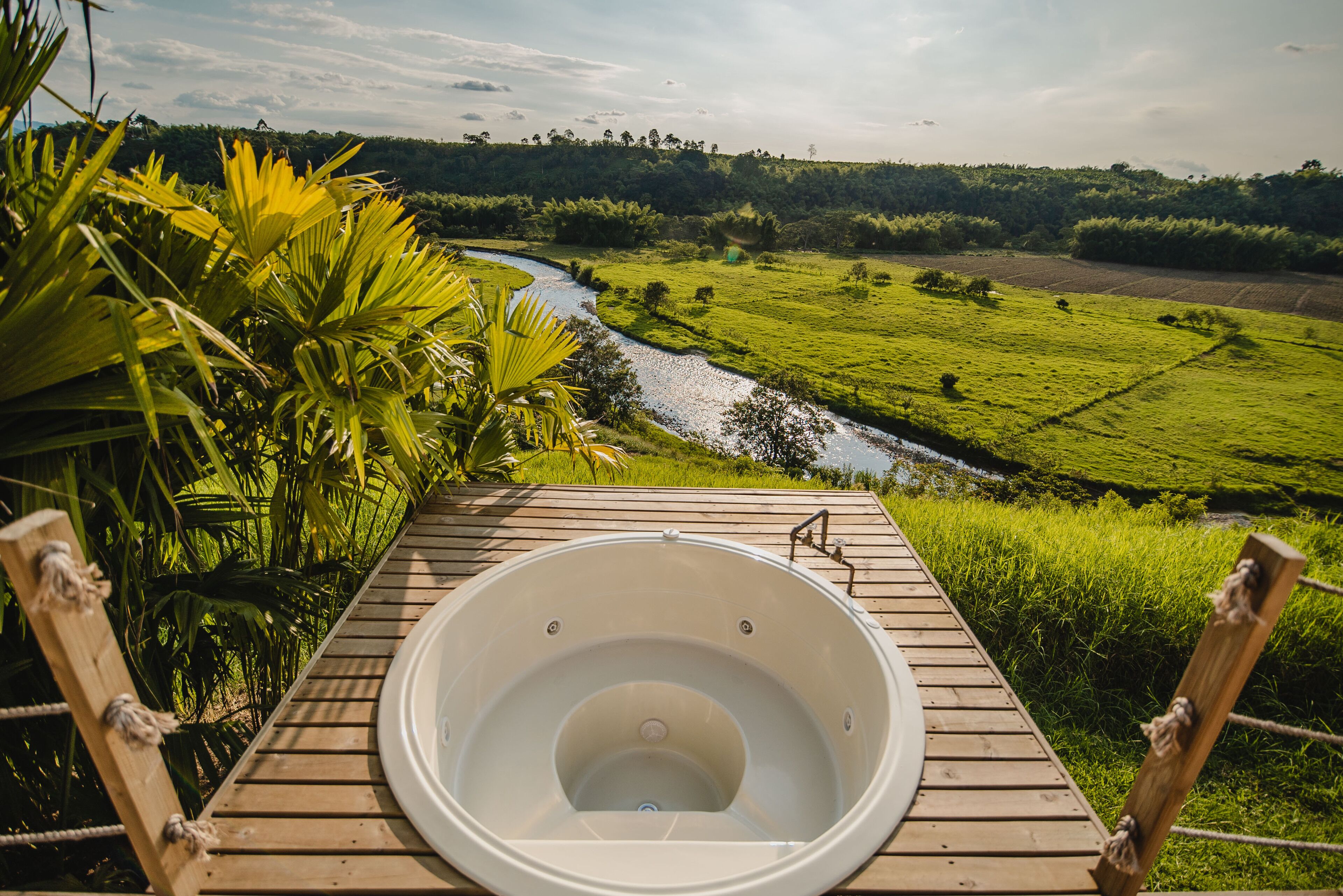 Un jacuzzi en una terraza de madera con vistas a un río y campos verdes exuberantes.