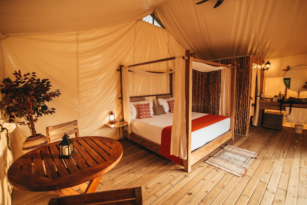 Una cama con dosel y cuatro columnas, una mesa de madera con un farol y una planta en maceta en una habitación cubierta.