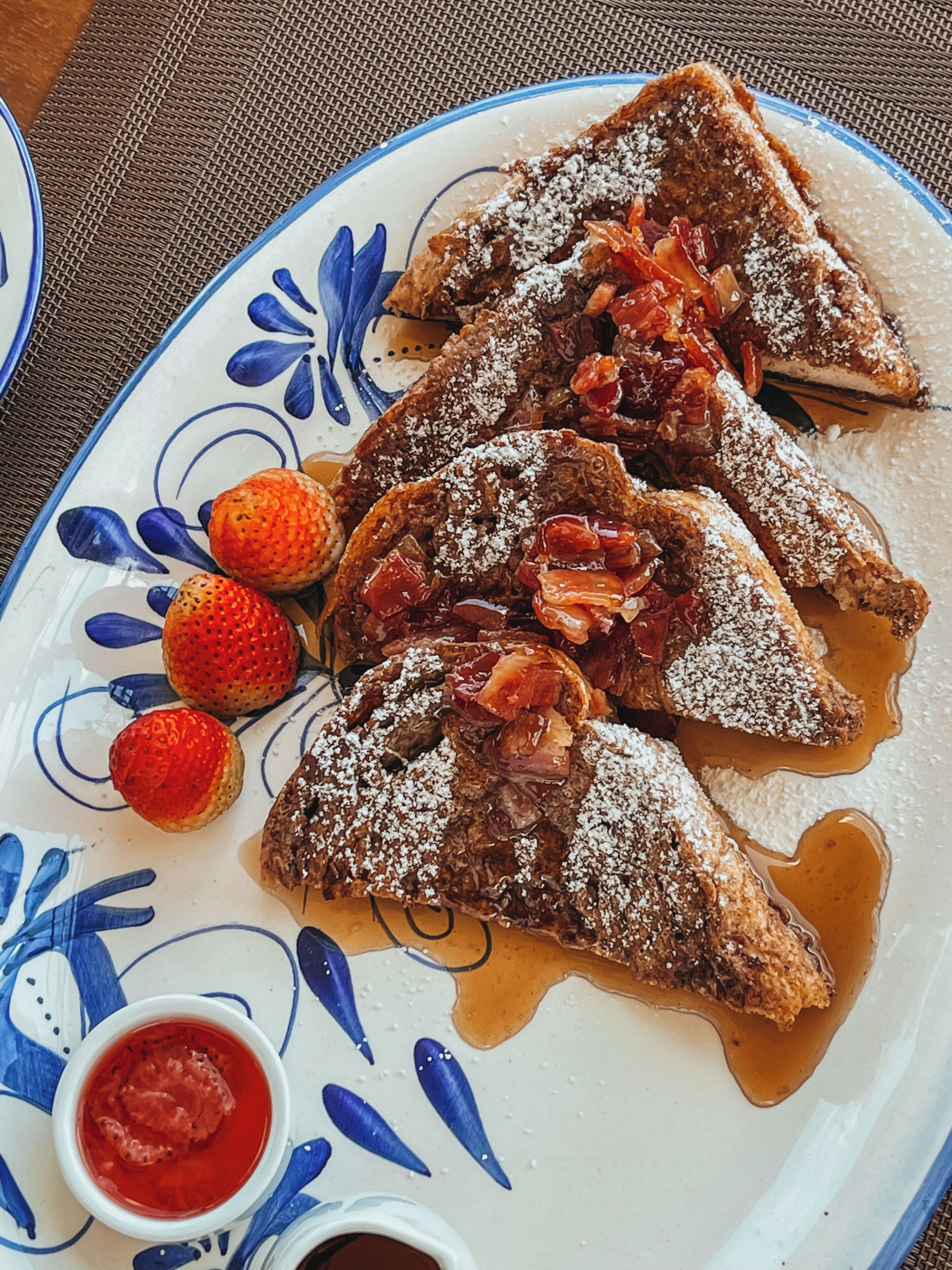 A plate of French toast with syrup and powdered sugar, accompanied by strawberries and a small bowl of red sauce.