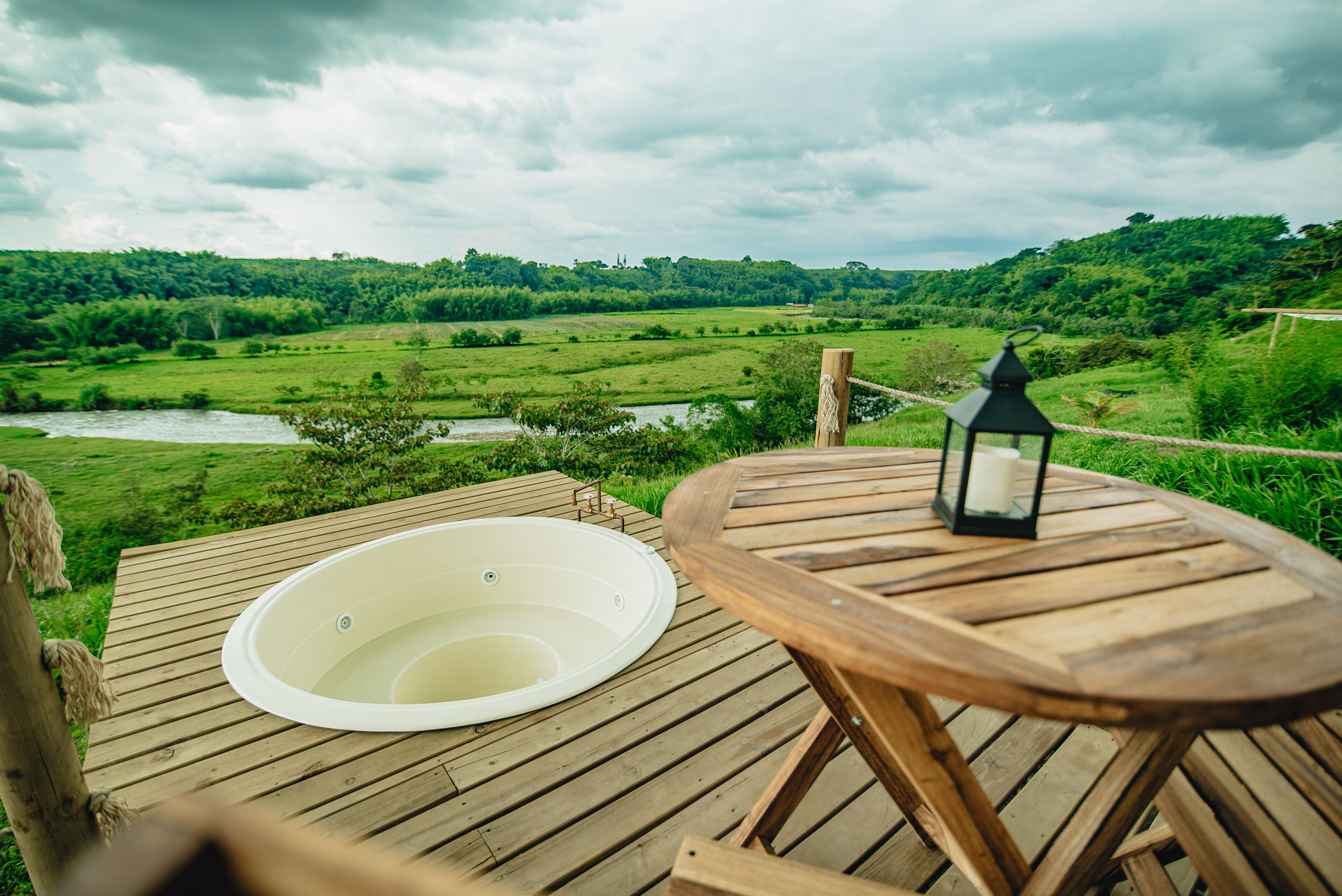 A wooden deck with a hot tub and a lantern, overlooking a lush green landscape with a river.