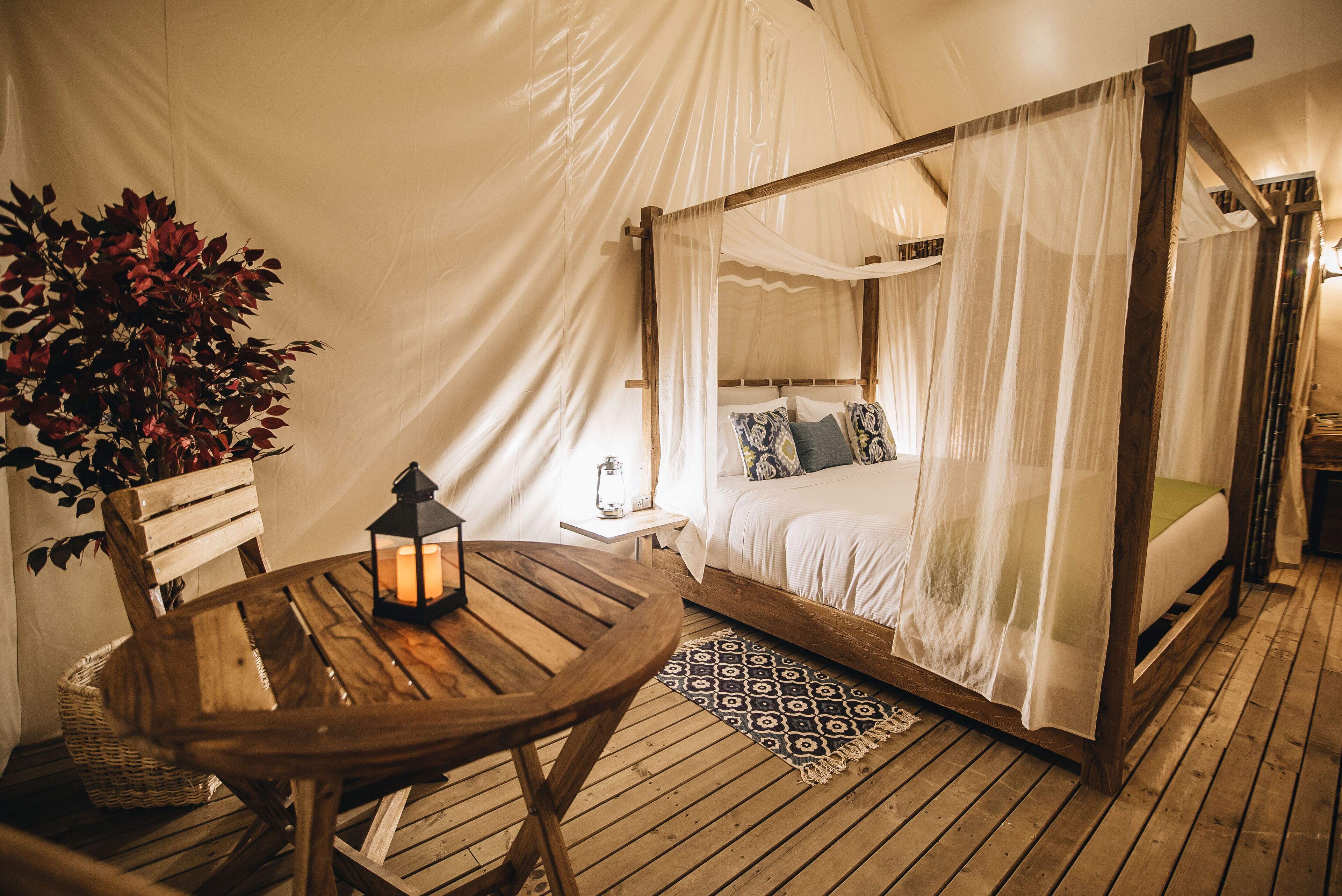 A tented room with a four-poster bed, a wooden table with a lantern, and a wicker basket.