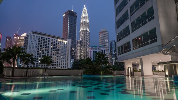 A swimming pool with a geometric pattern on the floor, surrounded by palm trees and modern buildings, including a prominent skyscraper with a spire.