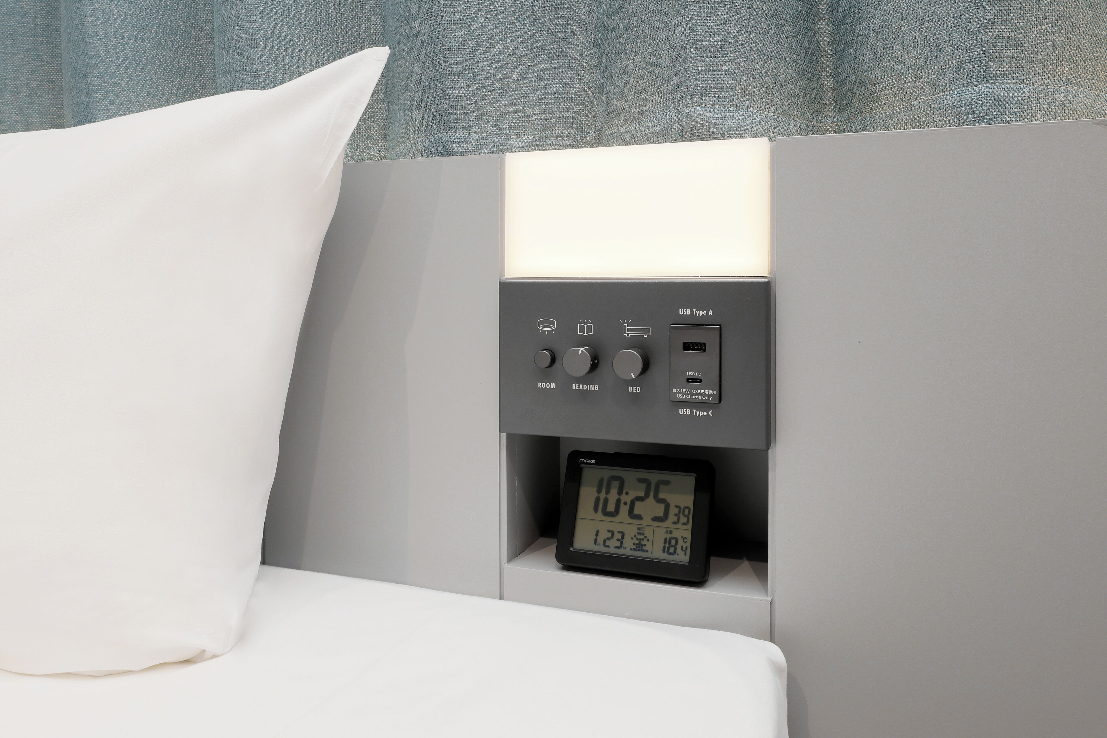 A bed with a white pillow, a digital clock displaying the time, and a control panel with USB ports and buttons labeled 'ROOM,' 'READING,' and 'BED.'