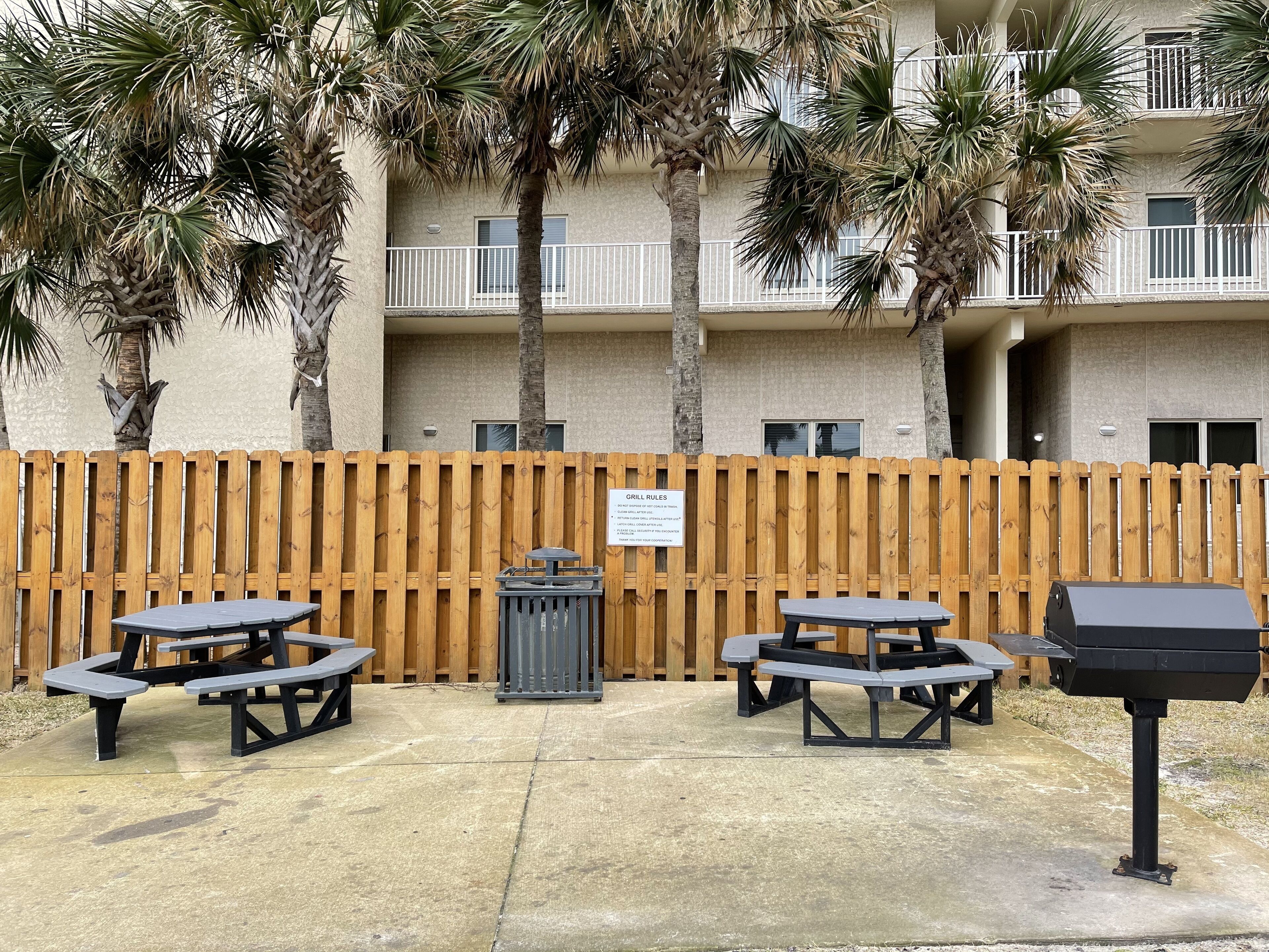 A patio area with picnic tables and a grill, surrounded by a wooden fence and palm trees, with a building in the background.