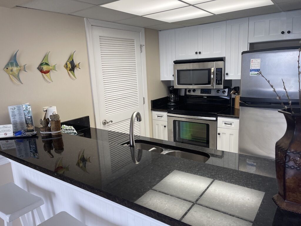 A kitchen with white cabinets, a stainless steel refrigerator, a microwave, and an oven. There are decorative fish on the wall and a vase on the counter.