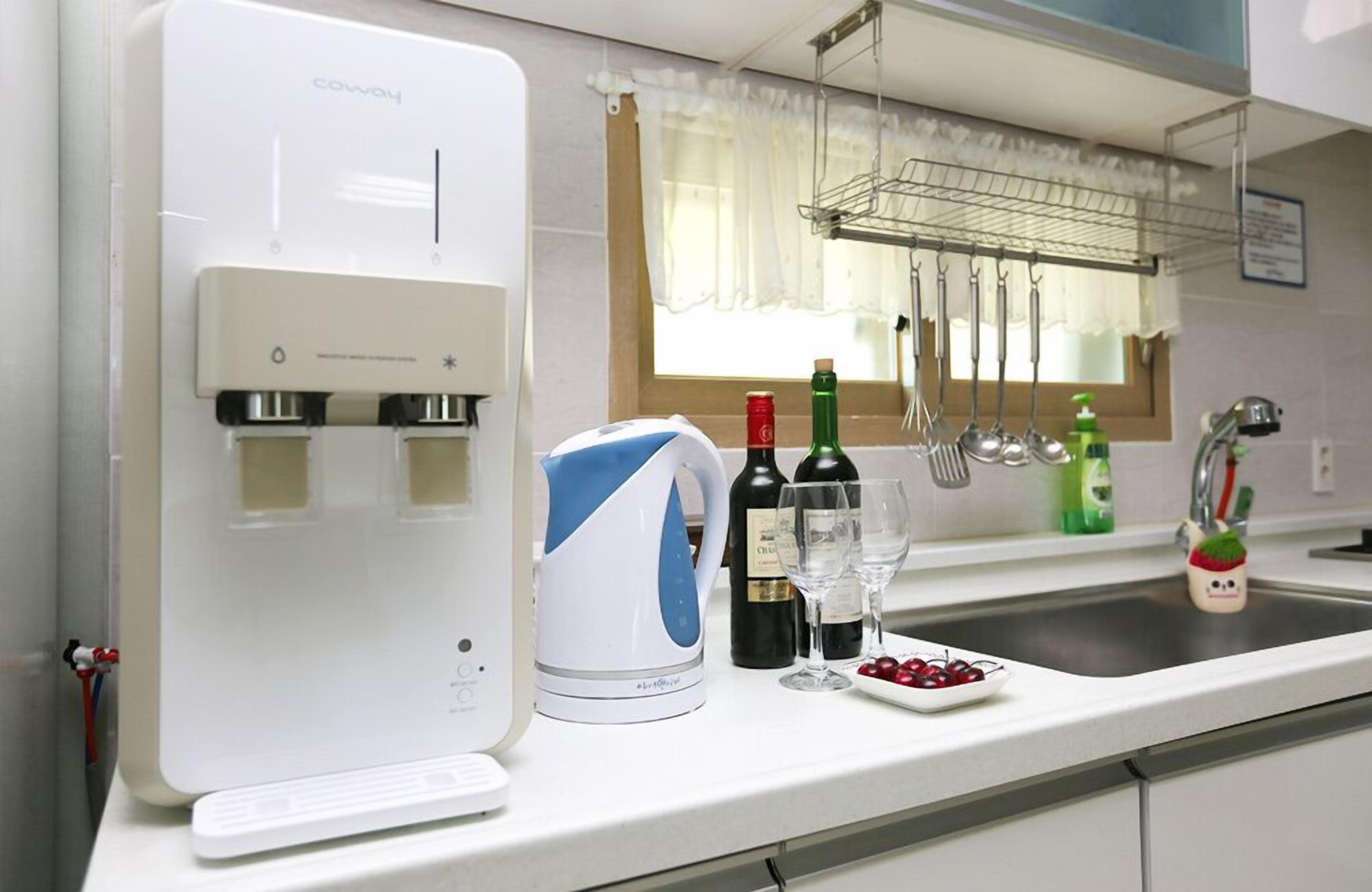 A kitchen with a water dispenser, a kettle, wine bottles, glasses, and a fruit plate.