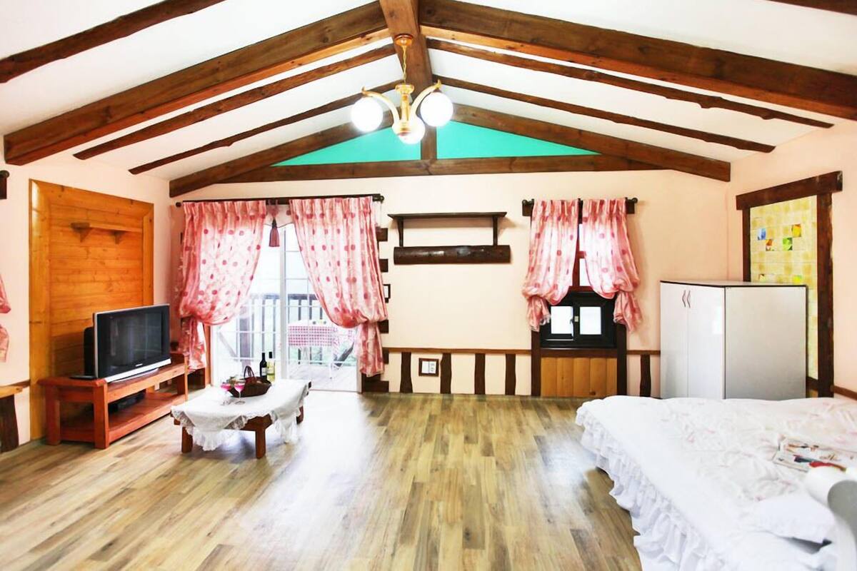 A cozy room with wooden beams, a television, a bed, a table with a tablecloth, and a refrigerator.