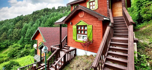 A wooden house with a red exterior and green shutters, a staircase leading to the entrance, and a view of a forested hillside. - Camp Star Pension (Jeongseon)