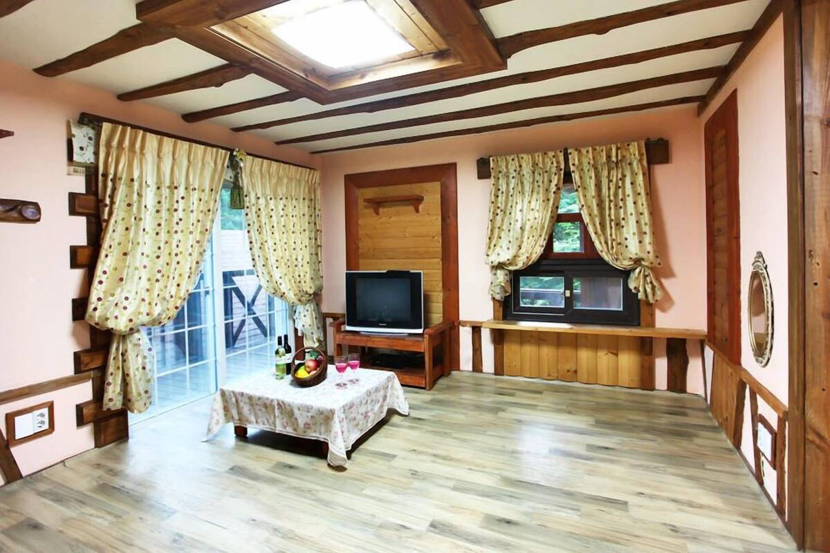 A cozy room with a wooden ceiling, a television on a wooden stand, a table with a fruit bowl, and curtains on the windows.
