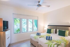 A hotel room with a bed, a TV, a ceiling fan, and a window with a view of greenery.