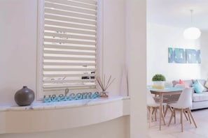 Une pièce avec une fenêtre, un vase, une pancarte « Bienvenue », une table avec des chaises et un canapé avec des coussins.