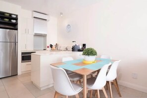 A modern kitchen with white cabinets, a stainless steel refrigerator, and a dining area with a wooden table and white chairs.