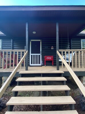 A wooden cabin with a porch, steps leading up to a door, and a Coca-Cola cooler on the porch.