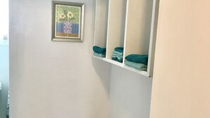Un coin de pièce avec une commode blanche, deux tableaux encadrés accrochés au mur et une étagère blanche où sont pliées des serviettes.