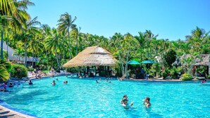 Piscina cercada por palmeiras e construções com telhados de palha.