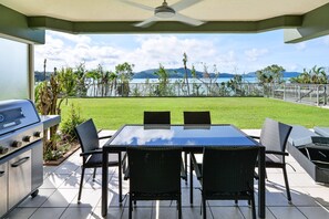 Un patio con una parrilla, una mesa de comedor y sillas, con vistas a un área verde y una masa de agua con montañas al fondo.