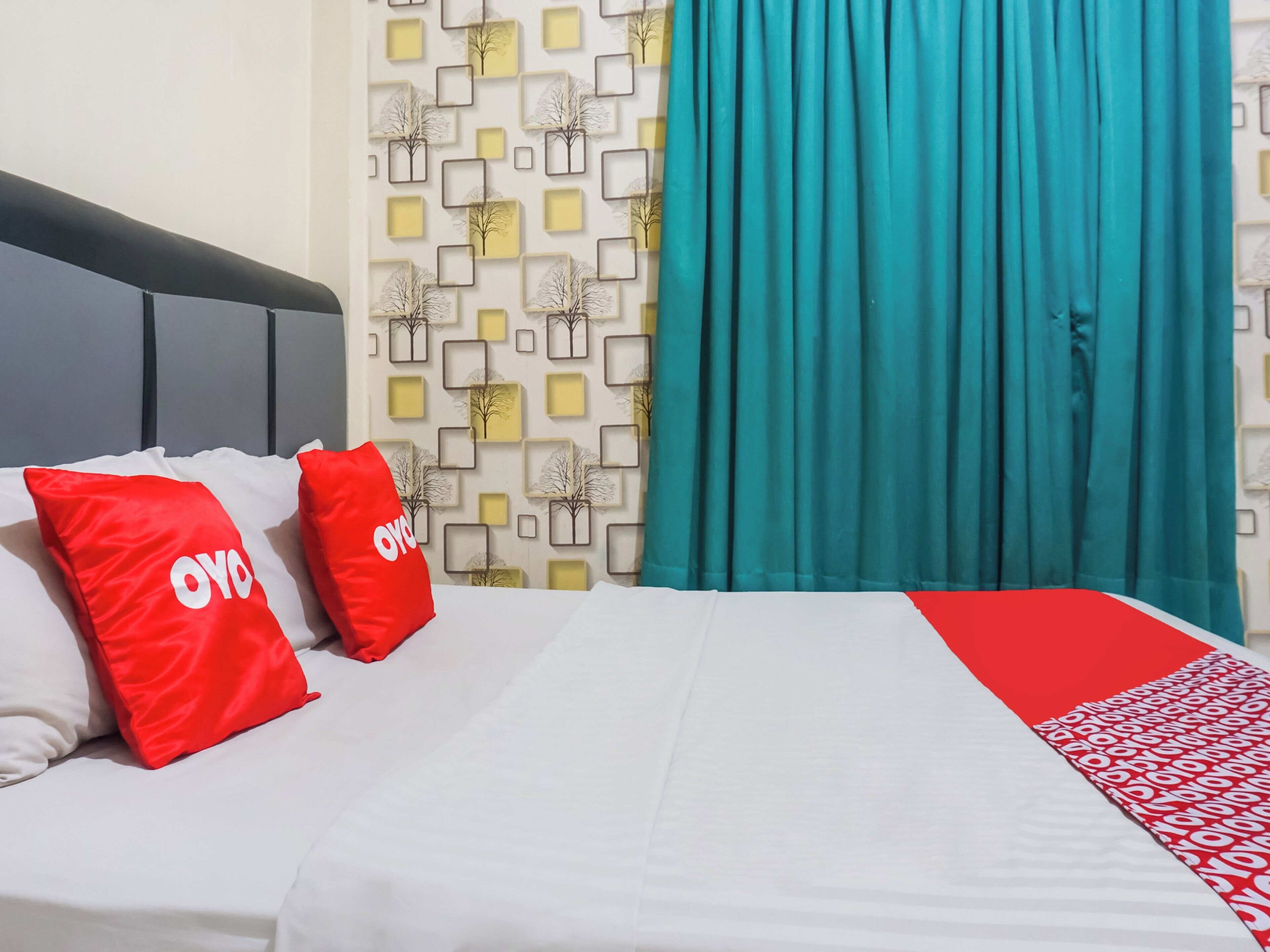 Hotelkamer met een bed, twee rode kussens met de tekst 'OYO', een patroonwand en tealblauwe gordijnen.
