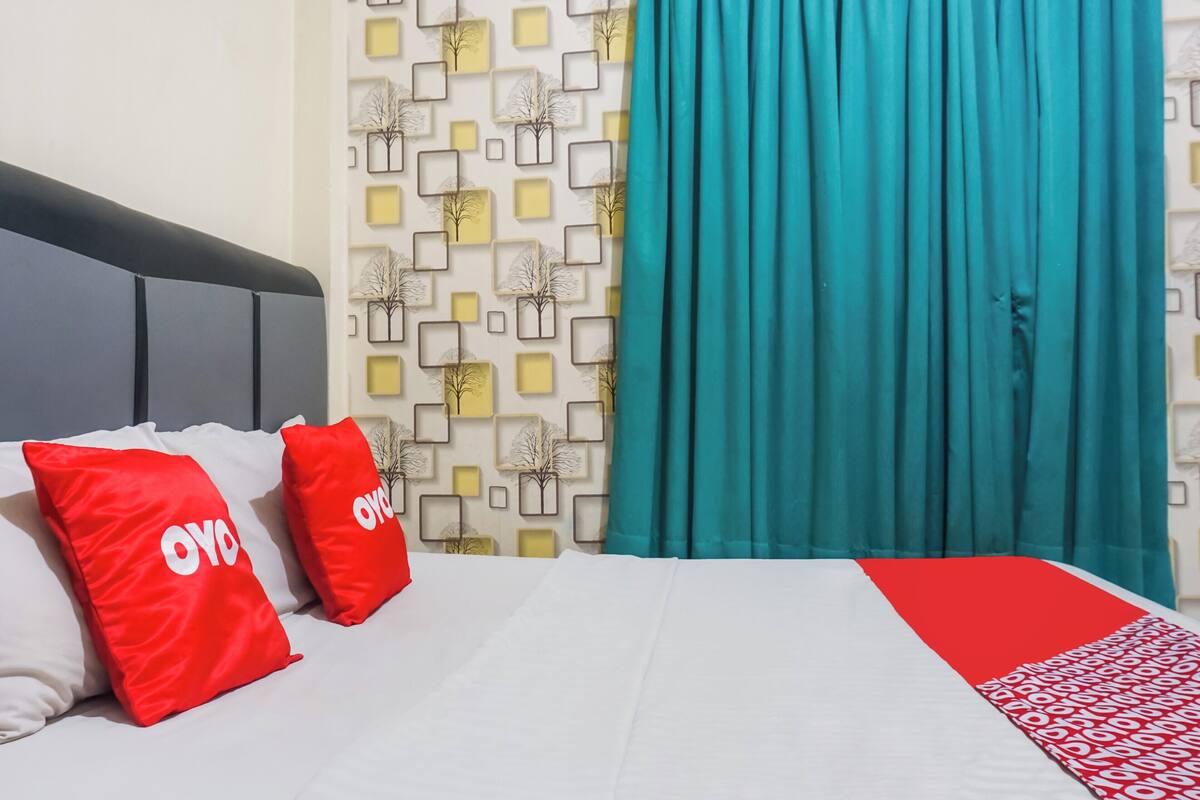 Ein Hotelzimmer mit einem Bett, zwei roten Kissen mit dem Schriftzug 'OYO', einer gemusterten Wand und türkisblauen Vorhängen.