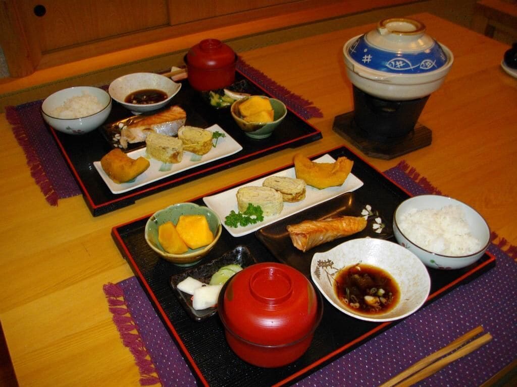 A traditional Japanese meal setup with various dishes, including rice, fish, and pickled vegetables, arranged on black trays with red and blue bowls.