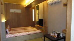 Een kleine kamer met een bed, een televisie, een tafel met dienblad en een airconditioner.