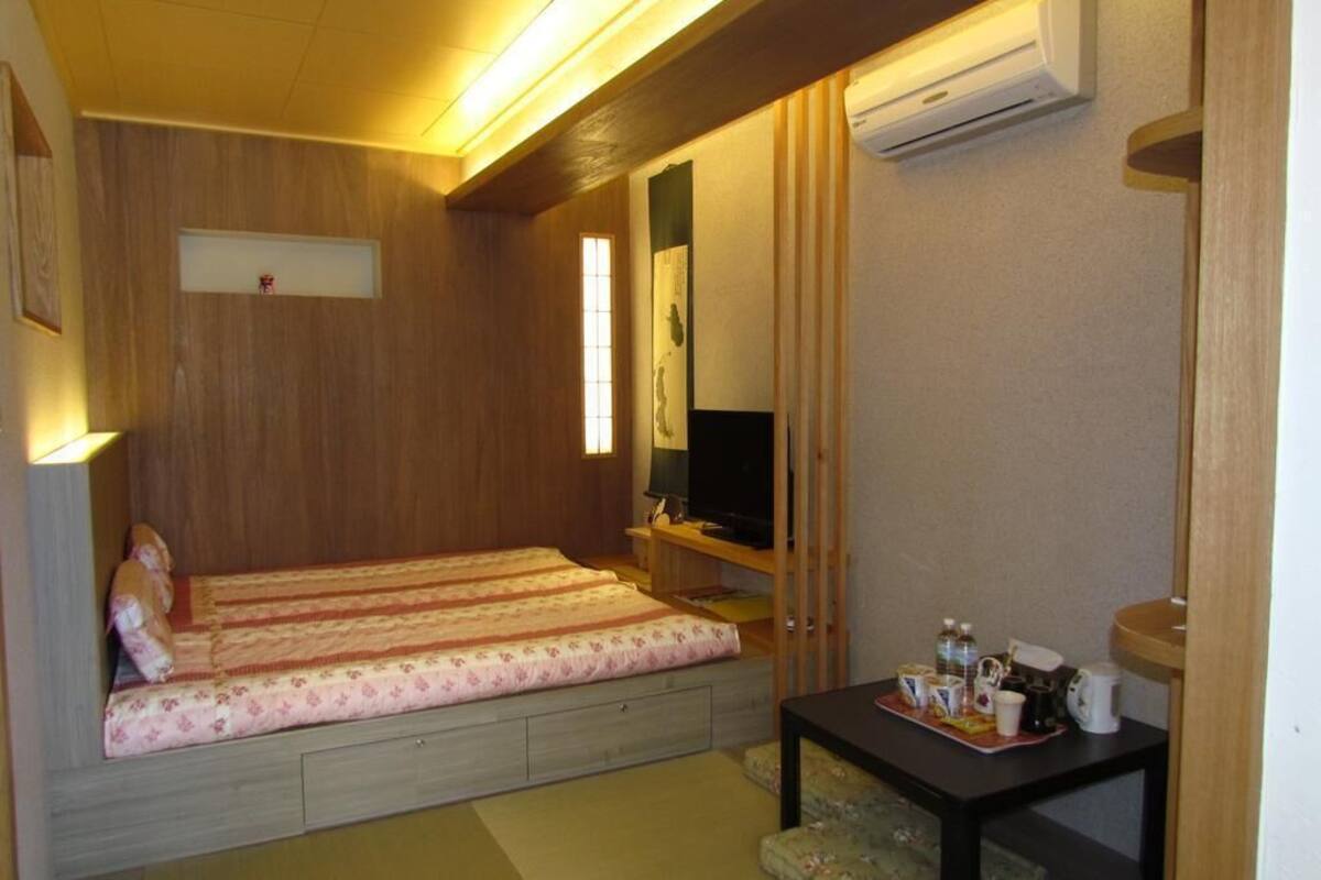 A small room with a bed, a TV, a table with a tray, and an air conditioning unit.