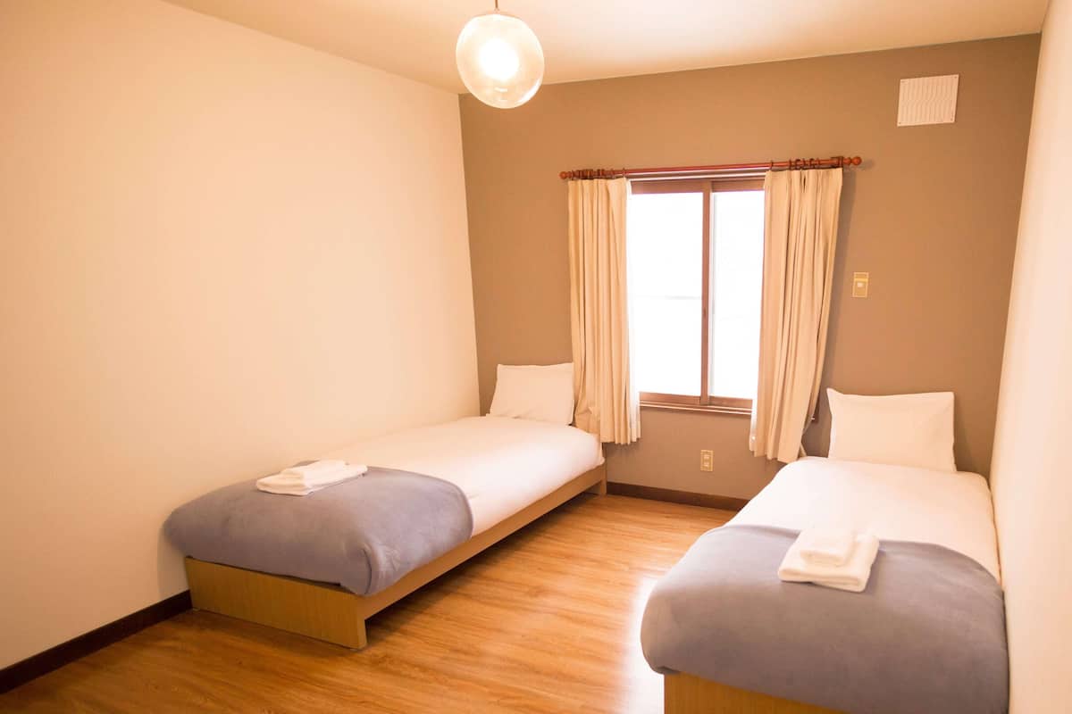 A room with two single beds, wooden flooring, a window with curtains, and a hanging light fixture.