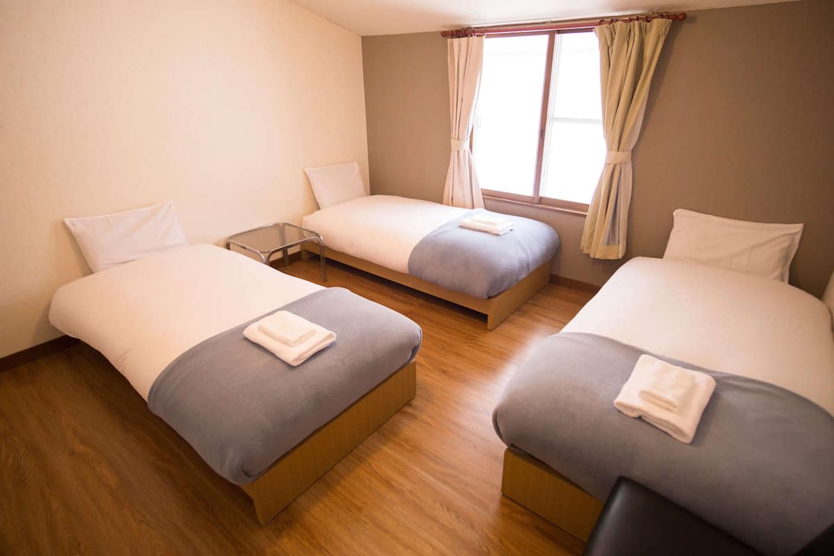 A hotel room with three single beds, wooden flooring, and a window with curtains.