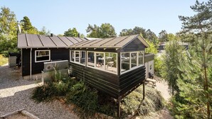 Eine kleine, einstöckige Holzhütte mit geneigtem Dach und großen Fenstern, umgeben von Bäumen und Pflanzen.
