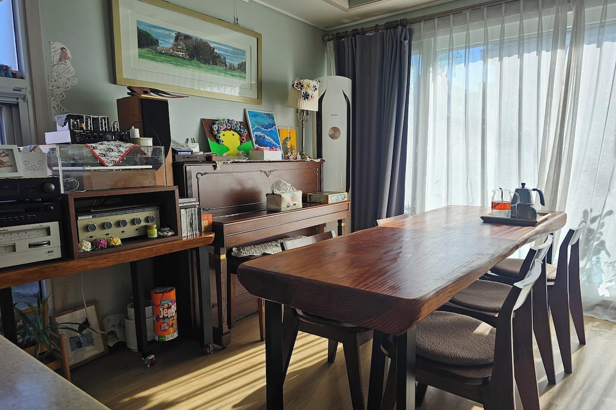 A room with a wooden table and chairs, a vintage stereo system, a framed picture on the wall, and a window with curtains.