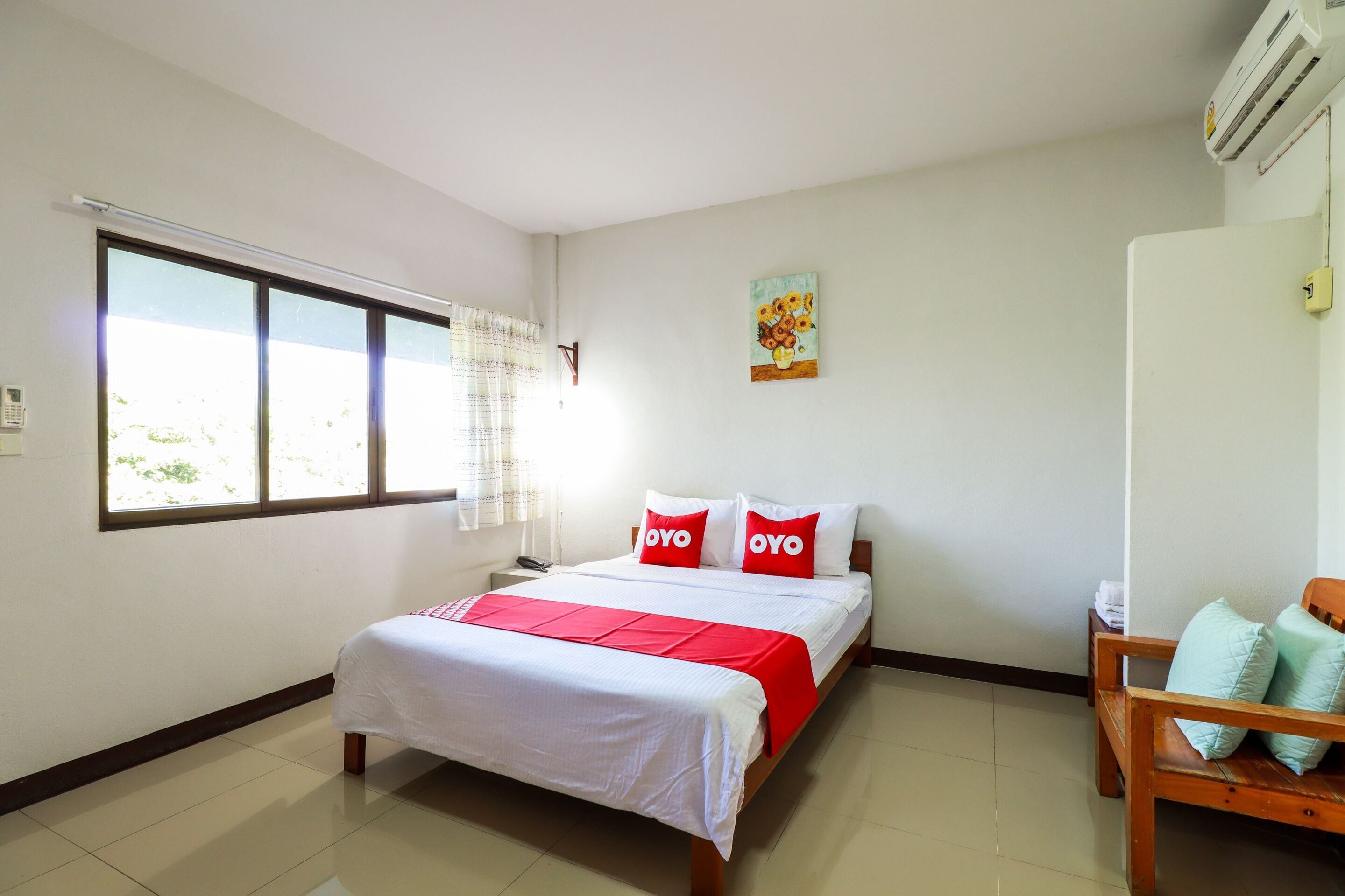 A hotel room with a bed, a wooden chair, a window with curtains, and a wall-mounted air conditioner.