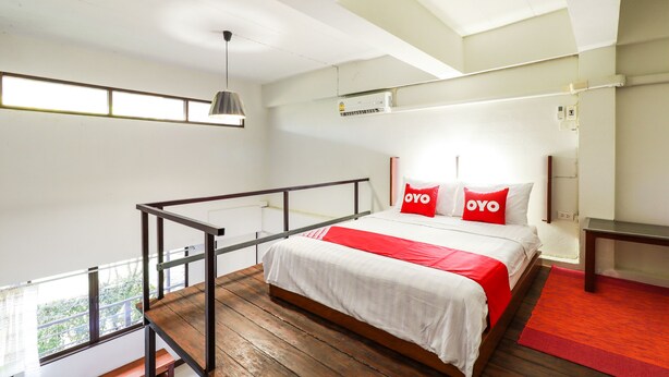 Une chambre moderne avec un plancher en bois, un lit avec une literie blanche et rouge, un oreiller rouge orné du texte « OYO », une suspension grise et une fenêtre offrant une vue.