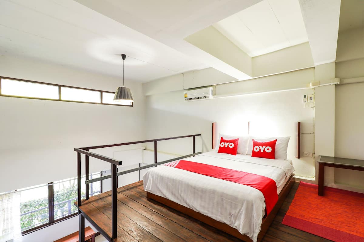 Une chambre moderne avec un plancher en bois, un lit avec une literie blanche et rouge, un oreiller rouge orné du texte « OYO », une suspension grise et une fenêtre offrant une vue.