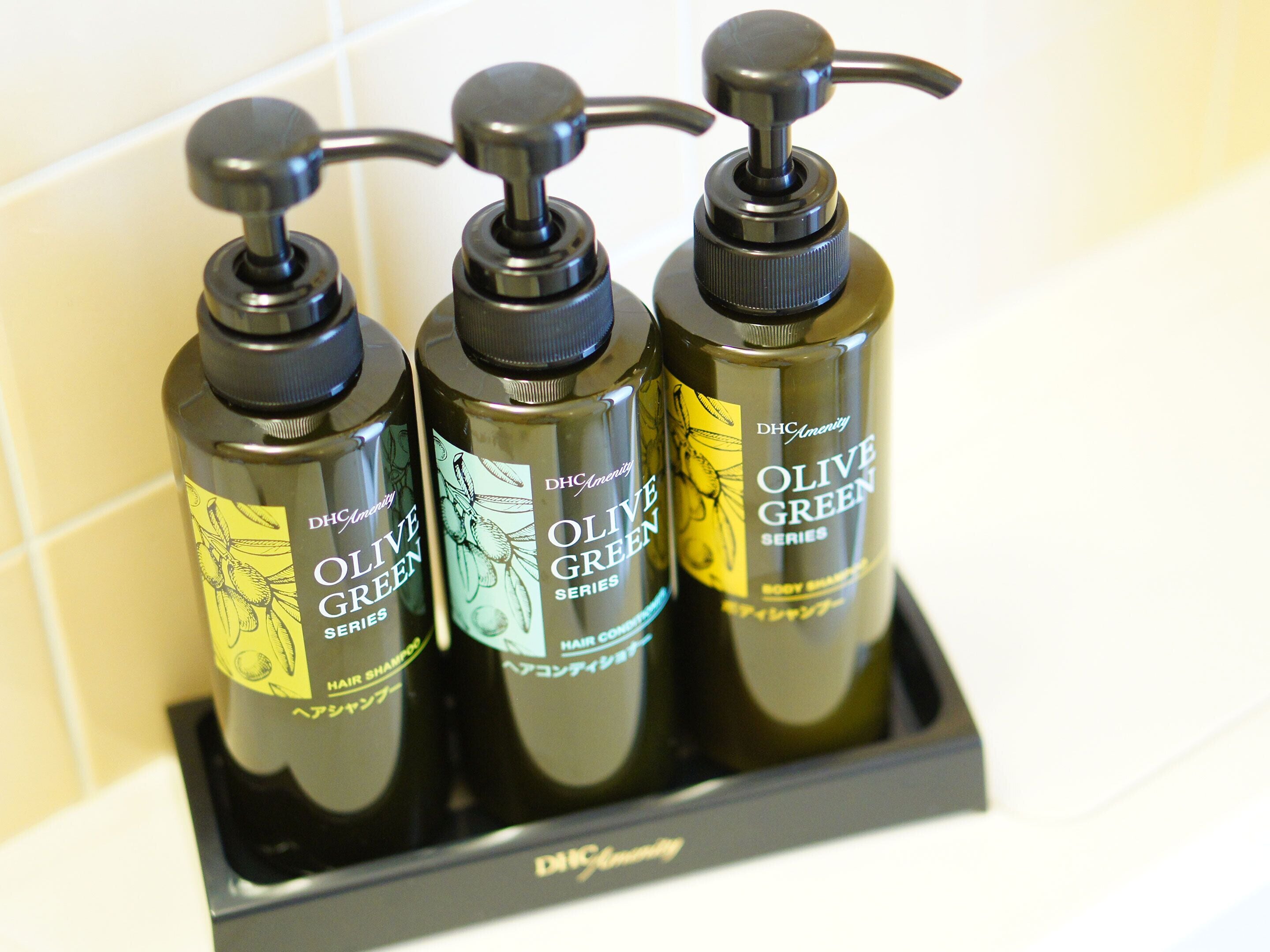 Three bottles of DHcAmenity Olive Green Series body shampoo in a black dispenser.