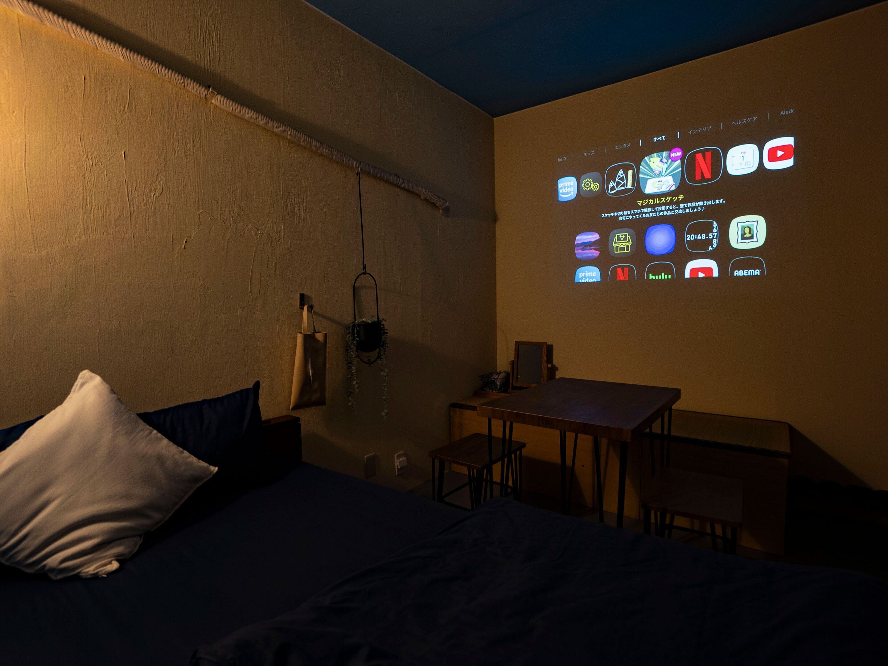 Um quarto com pouca luz, com uma cama, uma escrivaninha e uma projeção na parede exibindo diversos ícones de aplicativos.