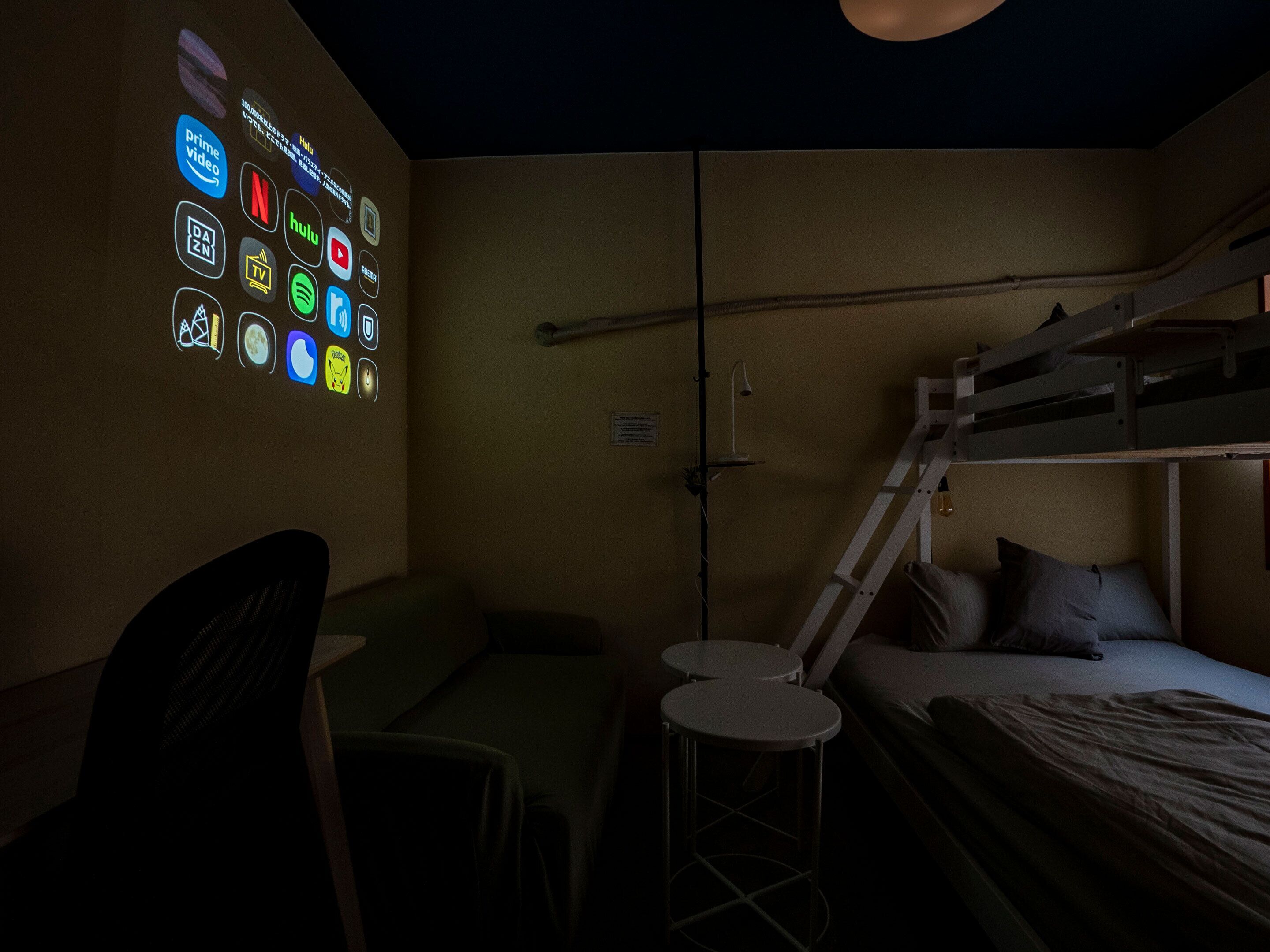 A dimly lit room with a bunk bed, a desk, and a chair. There is a wall with illuminated icons representing various streaming services.