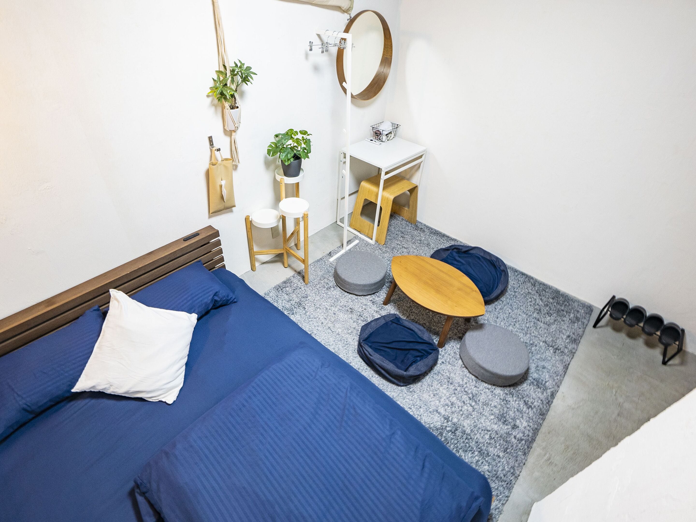 Un dormitorio compacto con cama, un taburete de madera, una mesita redonda y una mesita de noche blanca con espejo.