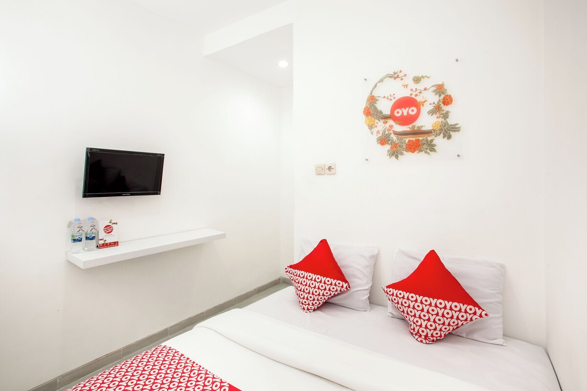 A hotel room with a bed, two pillows, a TV, a shelf with toiletries, and a wall decoration.